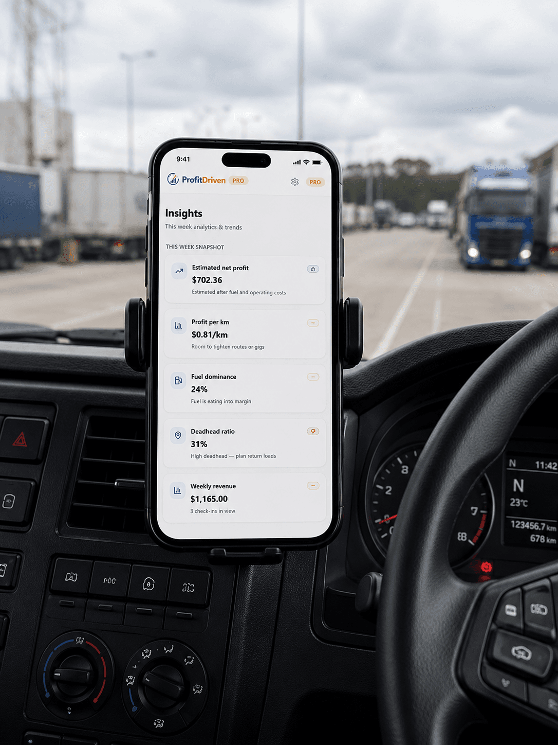 Smartphone mounted on truck dashboard displaying financial analytics app with semi-trucks in the background.