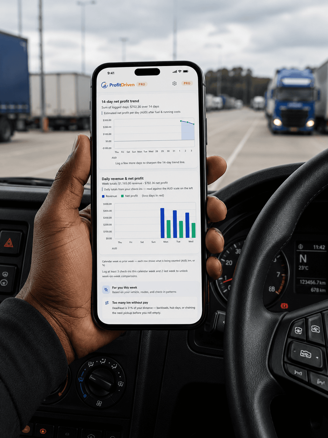 Hand holding a smartphone showing financial charts inside a truck cabin with trucks outside.