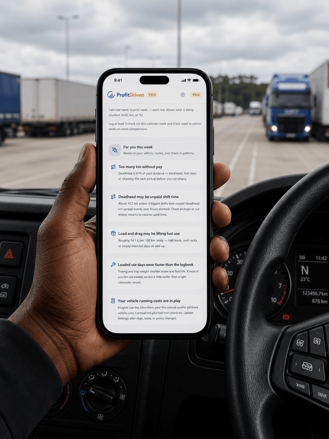 A hand holding a smartphone displaying a trucking logistics app inside a vehicle's driver cabin.